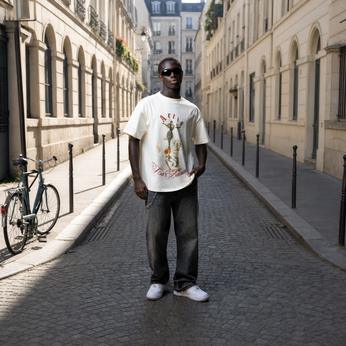 Man standing on a sunlit street wearing a white t-shirt with graphics and text.